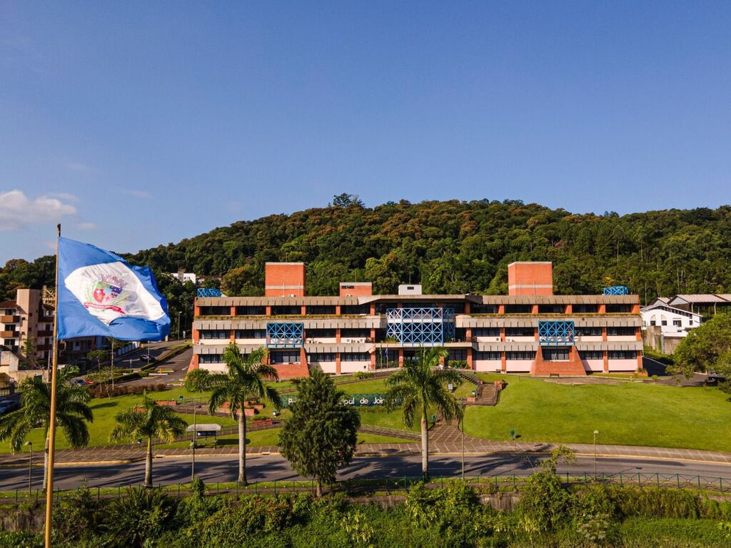 Esta foto apresenta uma vista aérea e ampla da Prefeitura Municipal de Joinville, localizada em Santa Catarina. A imagem destaca a arquitetura institucional integrada à paisagem natural da região.
